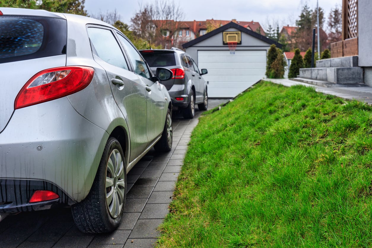Two cars are parked on a paved driveway next to a green grassy slope, with a white detached garage and basketball hoop visible in the background.