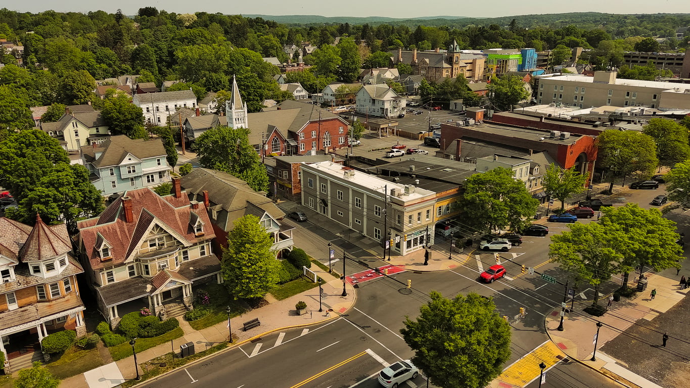 Aerial view of a small town with historic homes, shops, a white church, and tree-lined streets. Cars are parked along the roads and a few are driving through the intersection. Lush green trees fill the background.