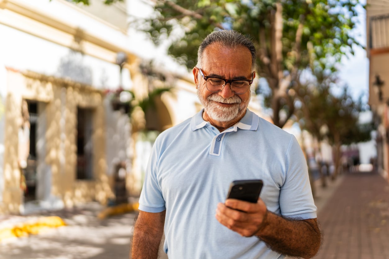 Smiling older man with glasses and a gray beard stands outdoors on a sunny day, looking at retirement tips on his smartphone. Trees and buildings line the background.