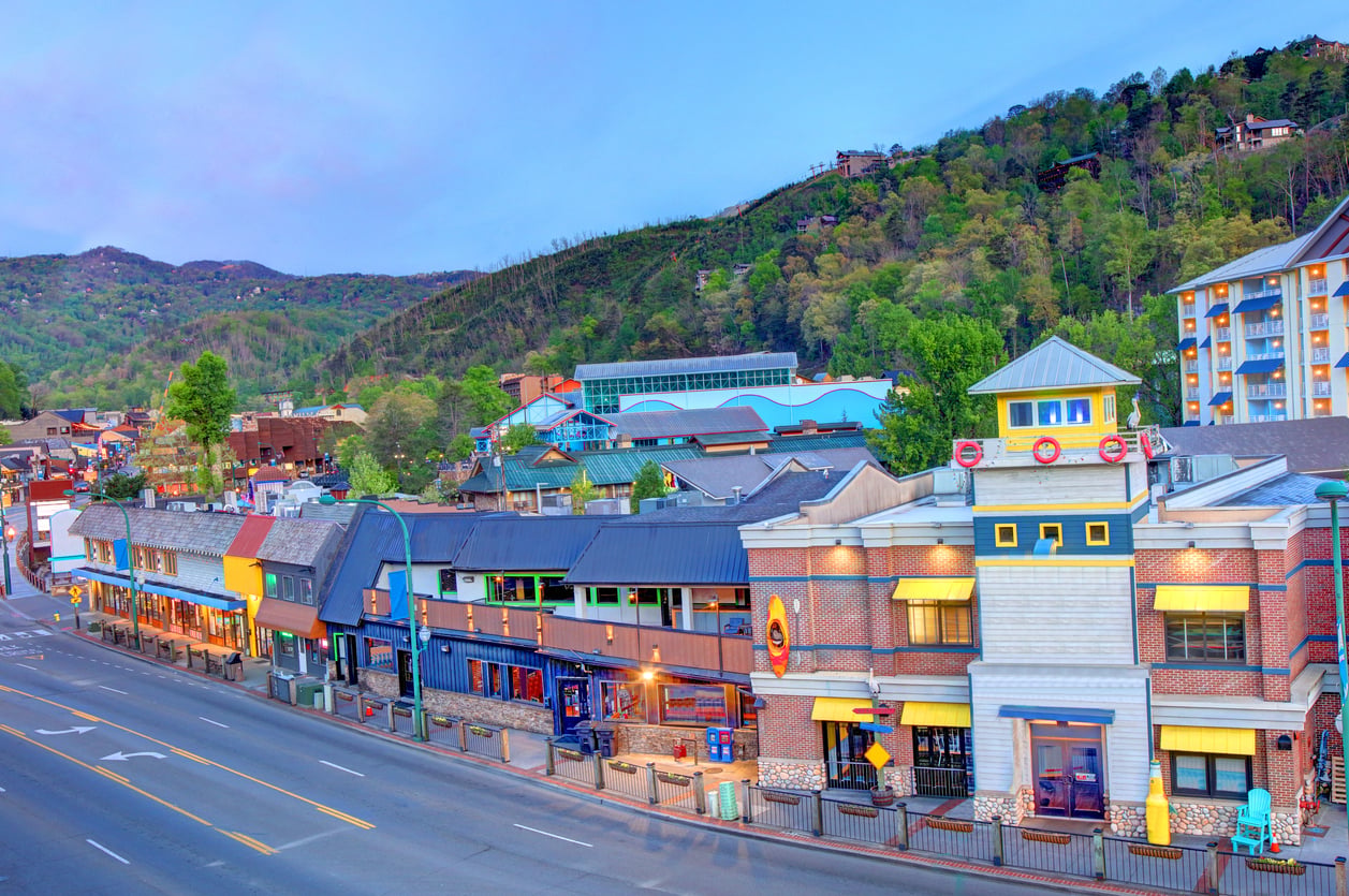 Colorful storefronts line a quiet street in a mountain town at dusk, with green hills in the background and lights glowing from shops and restaurants—though locals warn that, despite the inviting scene, some spots have a reputation for bad food.
