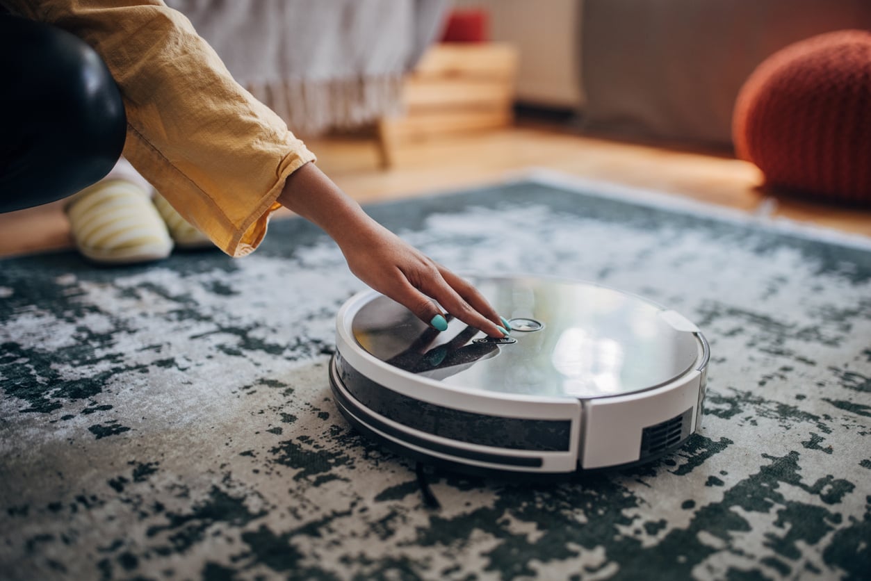 A person wearing a yellow sleeve presses a button on a white robotic vacuum cleaner as it sits on a patterned rug in a cozy room.