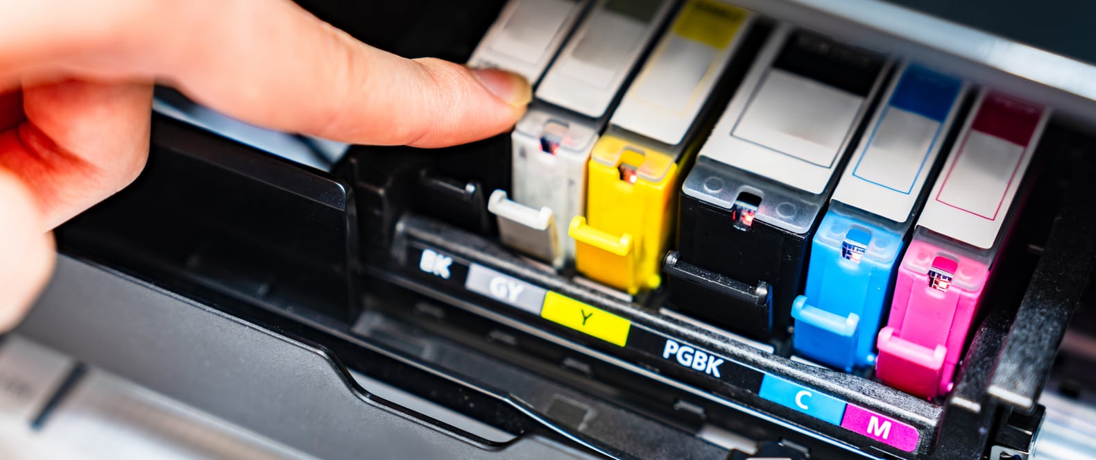 A person’s finger is touching a row of color ink cartridges inside a printer, with labels for black, gray, yellow, photo black, cyan, and magenta visible.