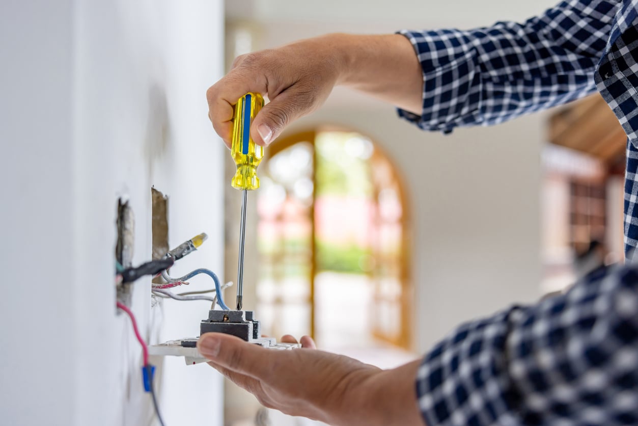 A person in a plaid shirt uses a yellow screwdriver to install or repair an electrical outlet on a white wall, with exposed wires visible and an open doorway in the blurred background.