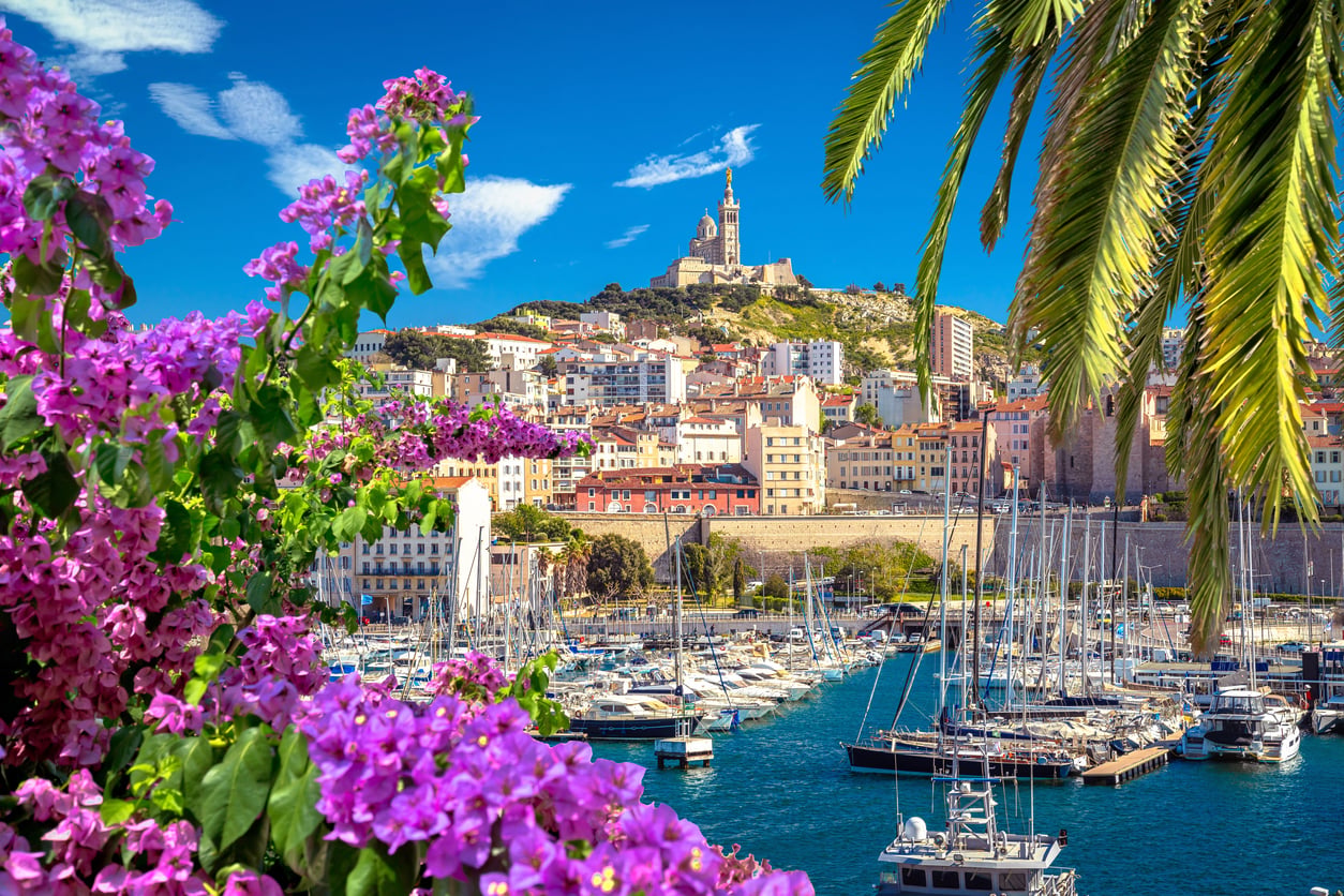 Colorful marina with sailboats, vibrant pink flowers, palm trees, and hillside buildings under blue sky; a church stands atop the hill in the background—a hidden gem among travel destinations boomers are skipping.