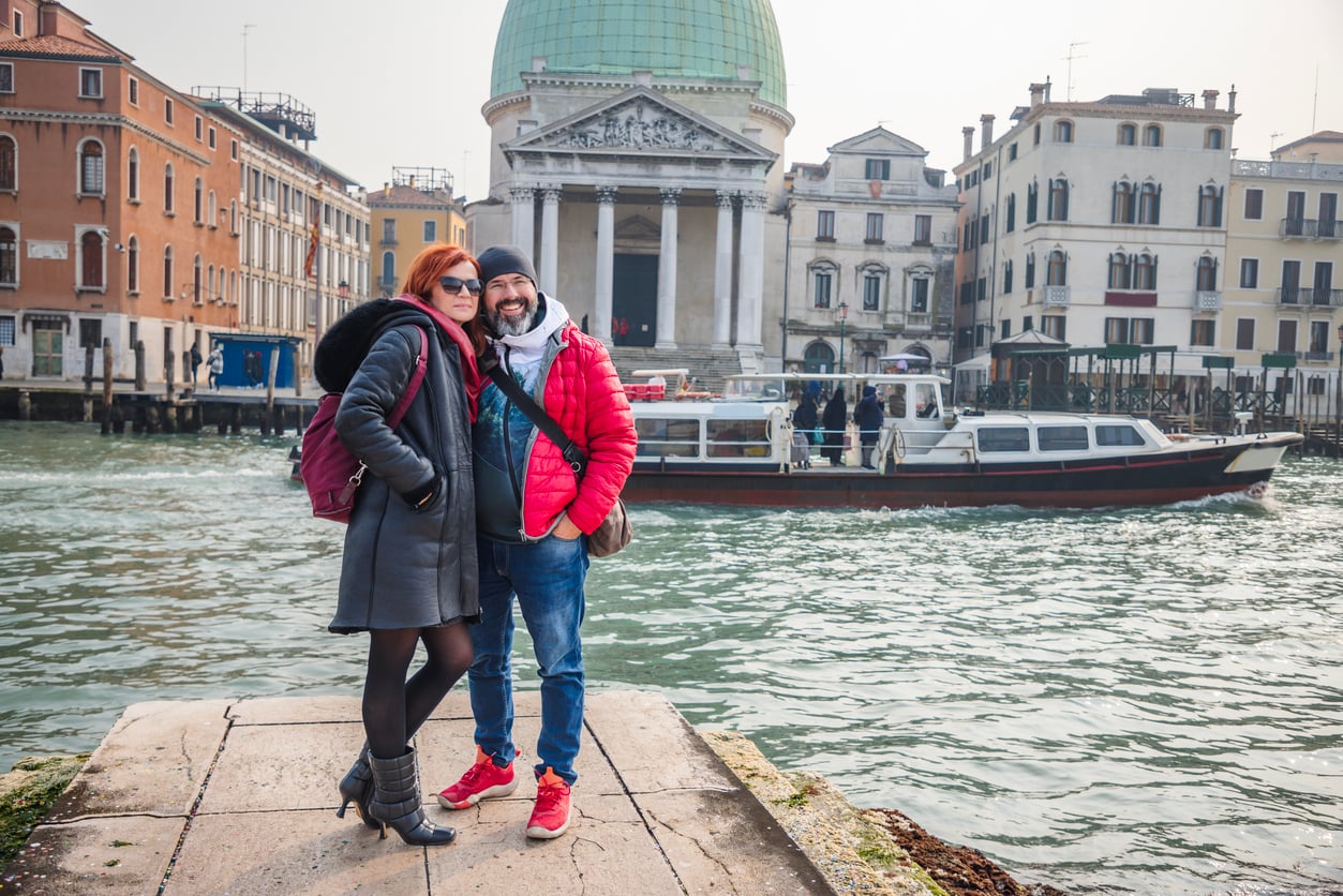 A smiling couple poses together by a canal in Venice, Italy—one of those classic travel destinations boomers are skipping these days—with historic buildings and a boat in the background. The woman wears a black coat and boots; the man wears a red jacket and sneakers.