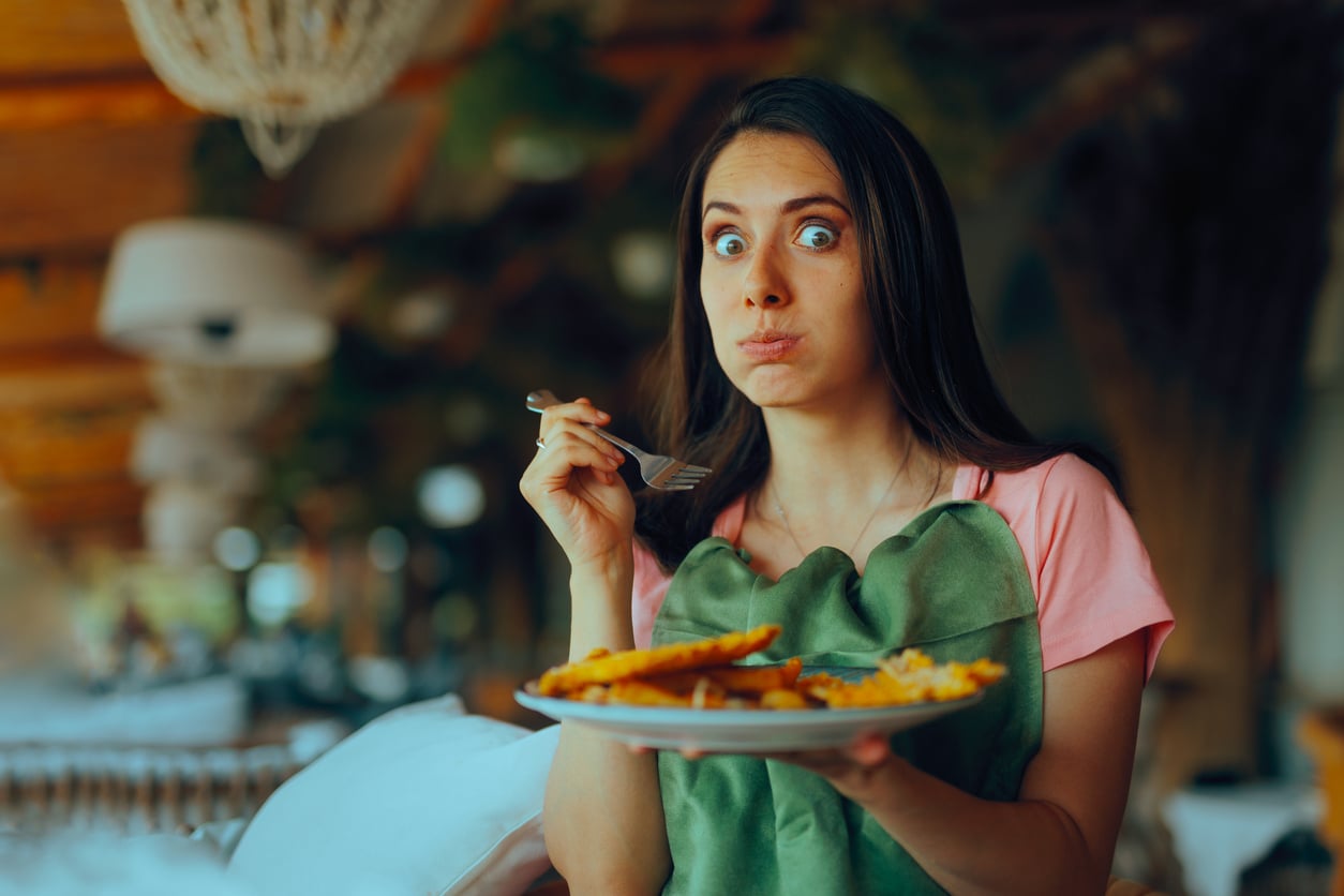 A surprised woman with wide eyes holds a fork and plate of food, cheeks puffed as if caught mid-bite, sitting in a restaurant with warm lighting and greenery in the background.