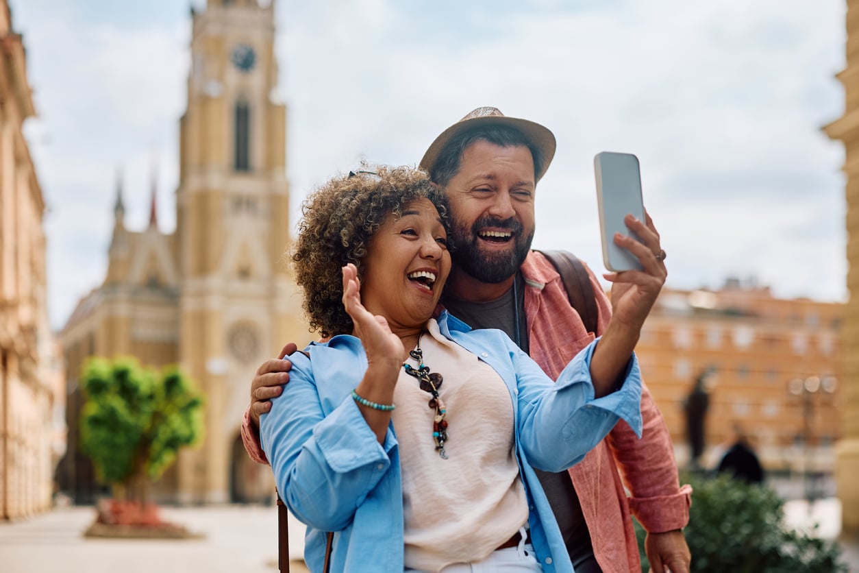 Smiling couple takes a selfie outdoors in a sunny city square, showcasing their joyful travel behaviors against the backdrop of historic buildings and a church tower. The woman waves while holding the phone, and both look happy and cheerful.