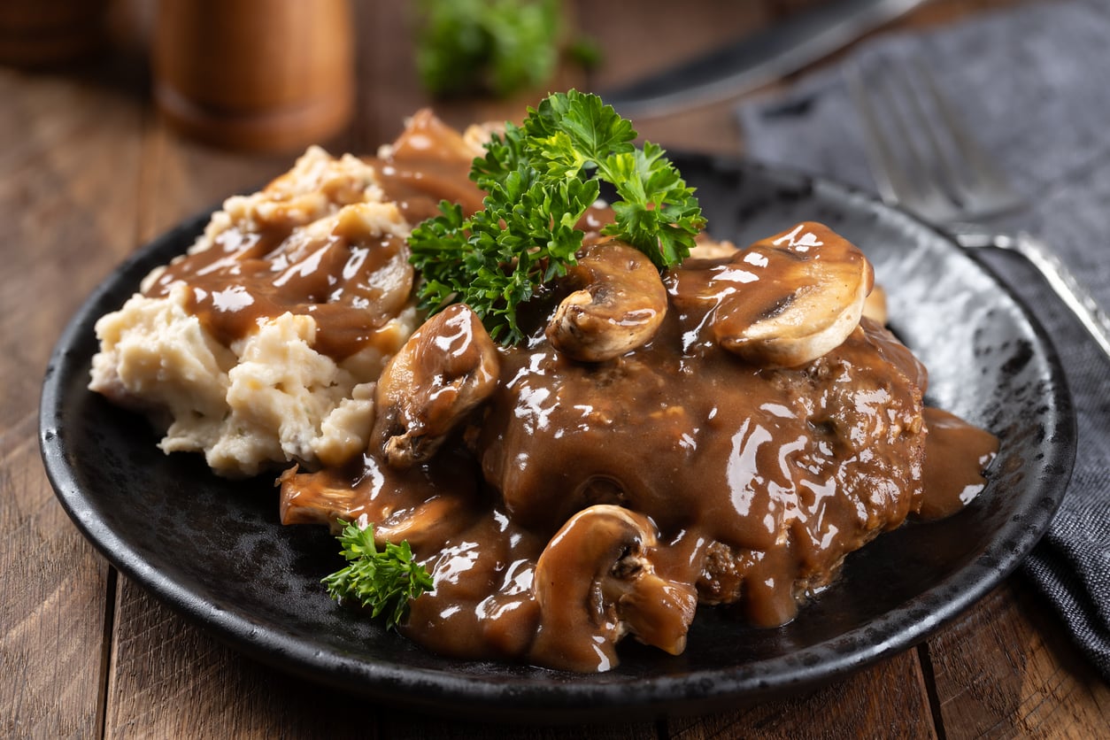 A plate of Salisbury steak with brown mushroom gravy, served with mashed potatoes and garnished with parsley on a dark plate—just like the classic meals middle-class moms made.