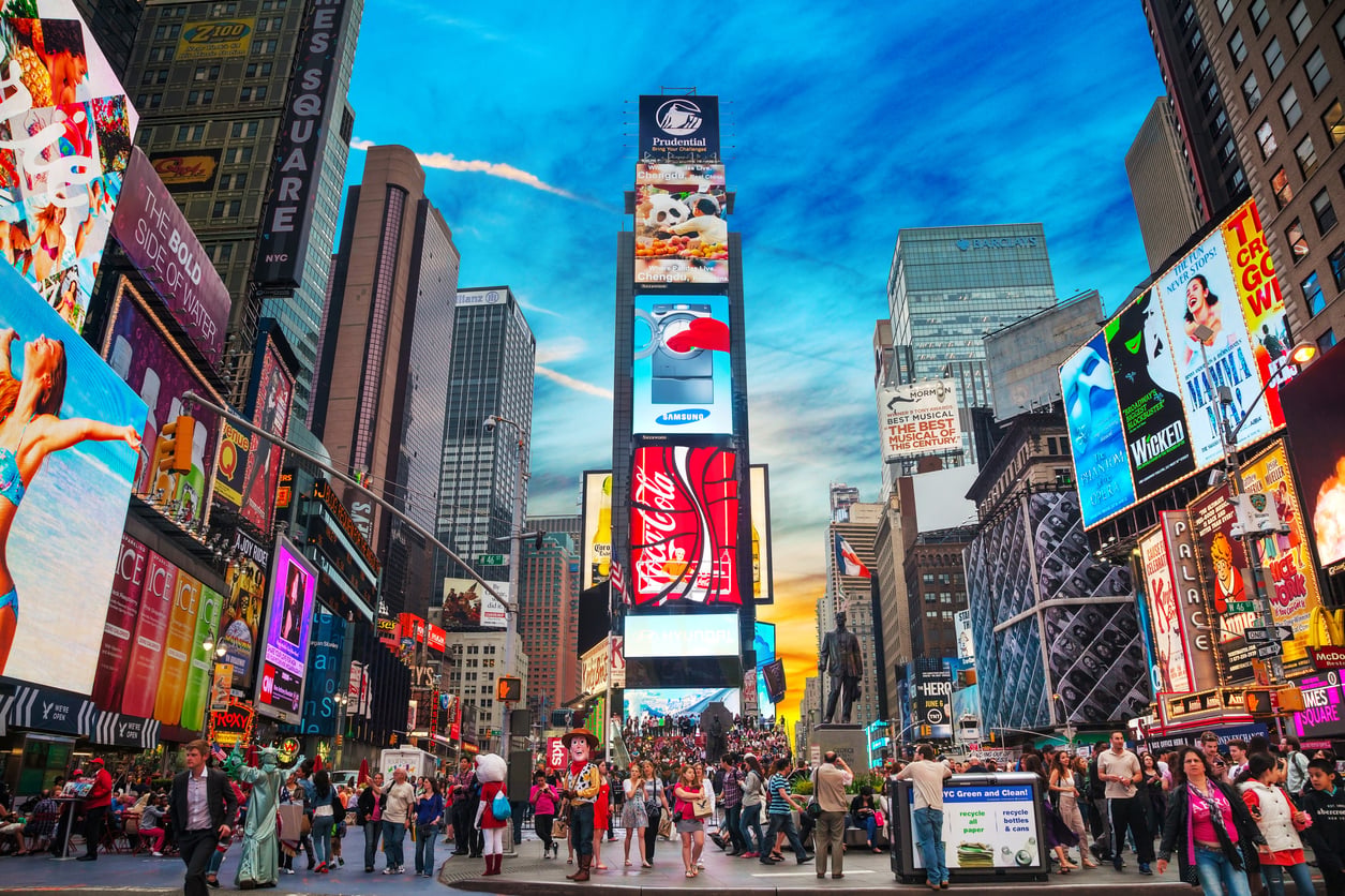 Crowds walk through Times Square in New York City, surrounded by tall buildings with bright digital billboards displaying colorful advertisements—including some for famously bad food—under a vivid blue and orange sky at sunset.