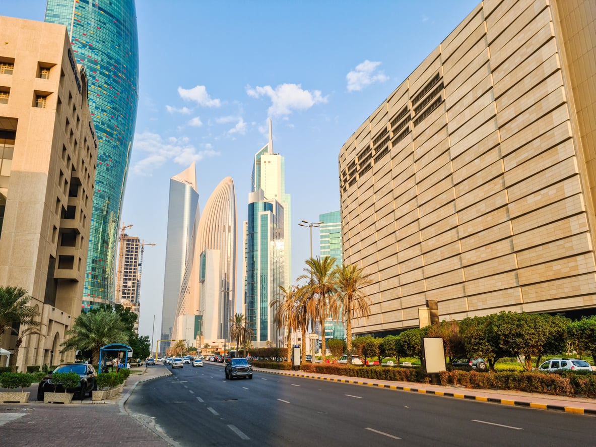 A city street in Kuwait with modern high-rise buildings, palm trees, parked cars, and a blue sky with scattered clouds offers a vibrant glimpse of daily life—be sure to stay updated on current Travel Restrictions And Advisories before planning your visit.