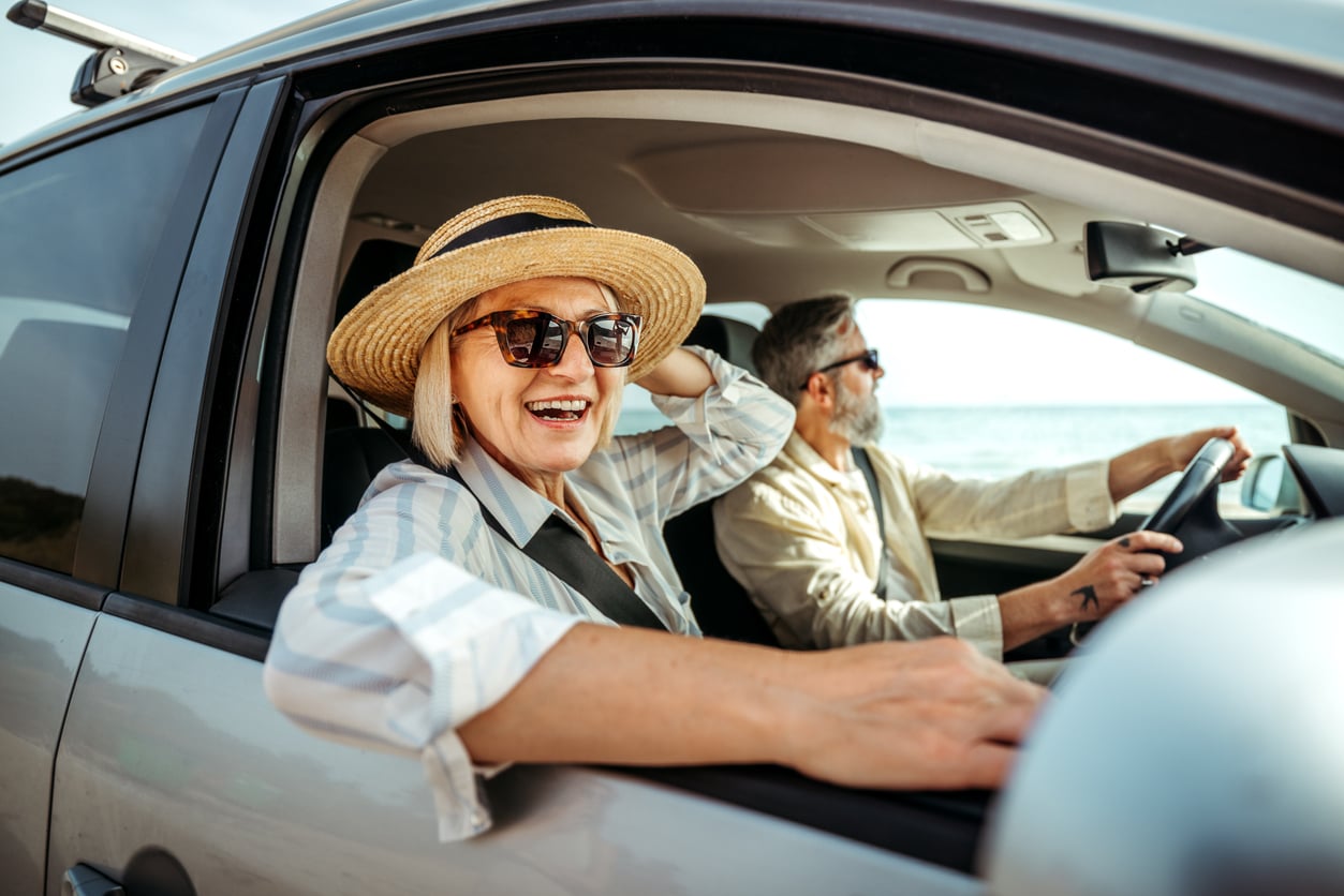 An older woman in sunglasses and a straw hat smiles from the passenger seat of a car, while a man drives. They appear to be enjoying a sunny day, possibly on a road trip or vacation near the coast.