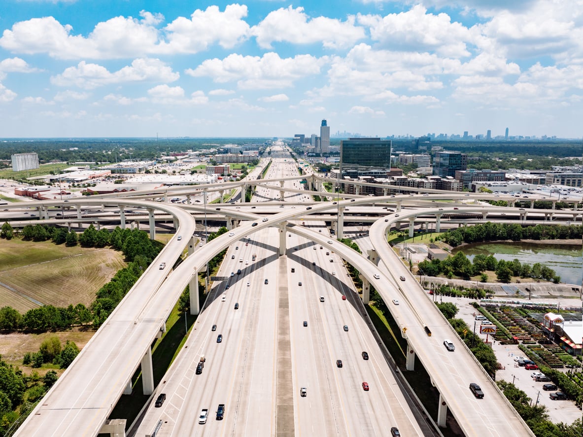 Aerial view of a large multi-level highway interchange with several overpasses and light traffic, surrounded by buildings, greenery, and a distant city skyline under a partly cloudy sky—a striking scene that perfectly illustrates one of the unique travel facts about the United States: its vast and intricate freeway systems connecting bustling cities with scenic landscapes.