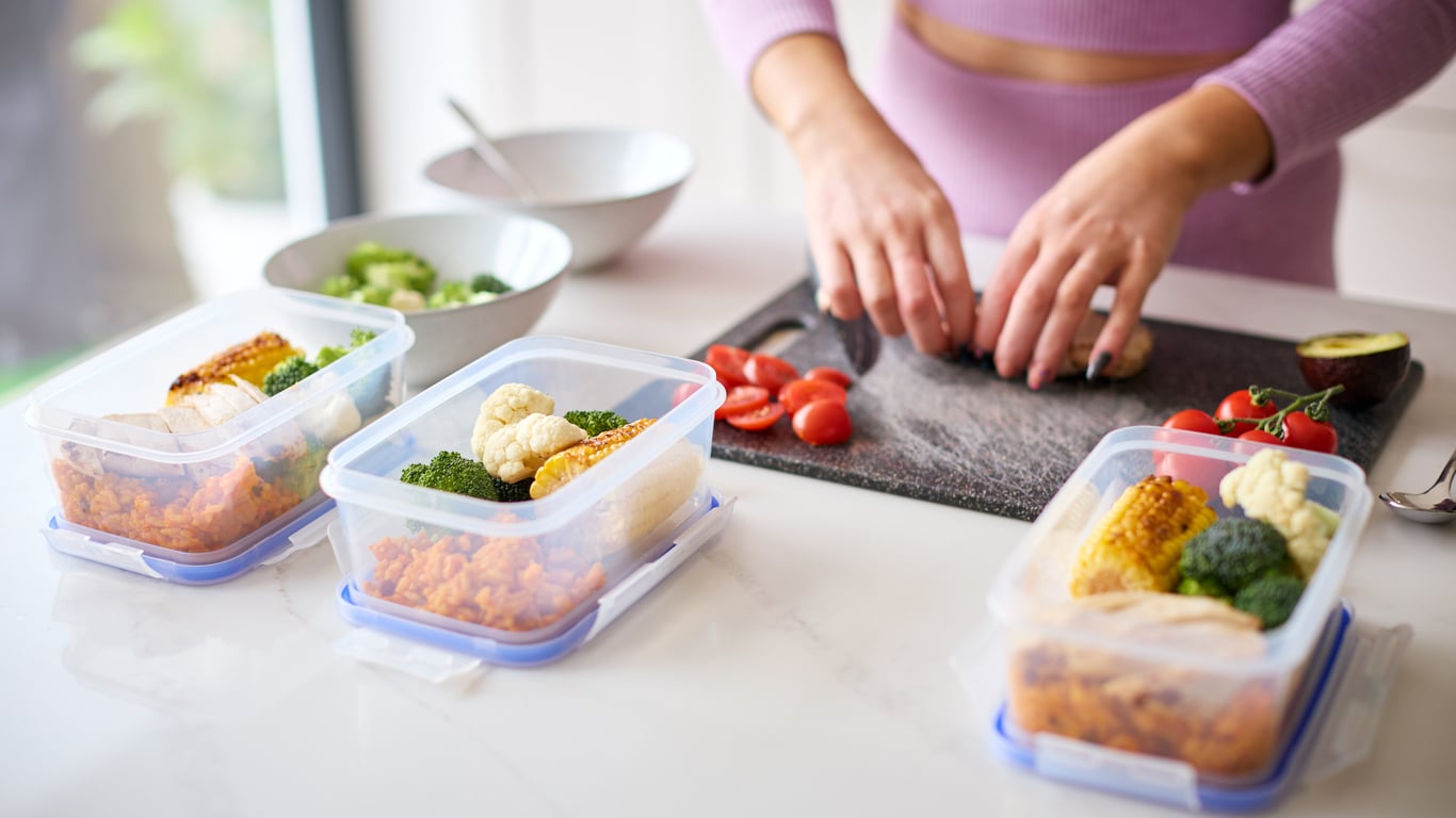 A person prepares meals on a kitchen counter, cutting tomatoes on a board. Three meal prep containers with rice, chicken, and vegetables are in the foreground.