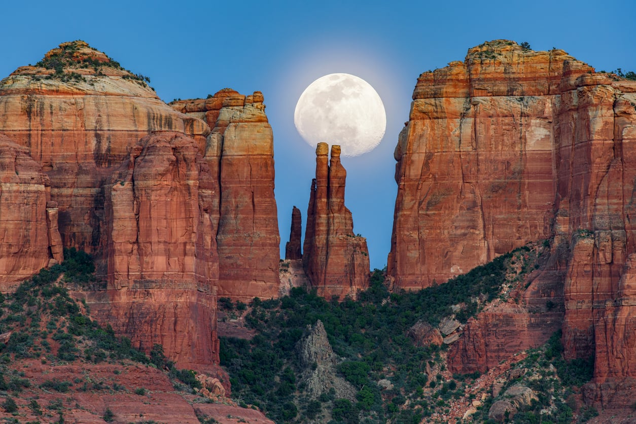 The full moon rises between tall, red rock formations with rugged textures and green shrubs at the base under a clear blue sky, offering a serene landscape that inspires adventure even after age 50.
