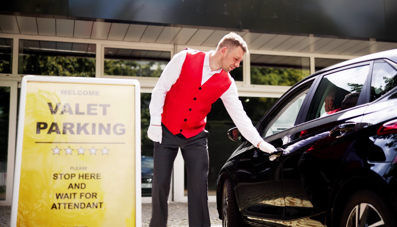 A valet in a red vest opens the door of a black car near a yellow “Valet Parking” sign outside a modern building.