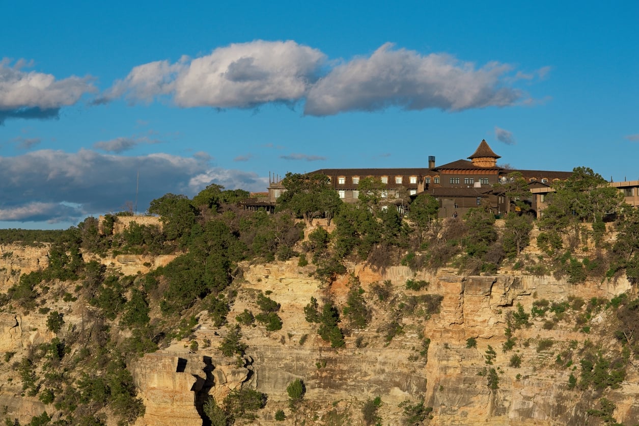 A historic lodge, part of the revered Historic Hotels collection, sits atop a rocky canyon rim, surrounded by trees under a blue sky with scattered clouds.