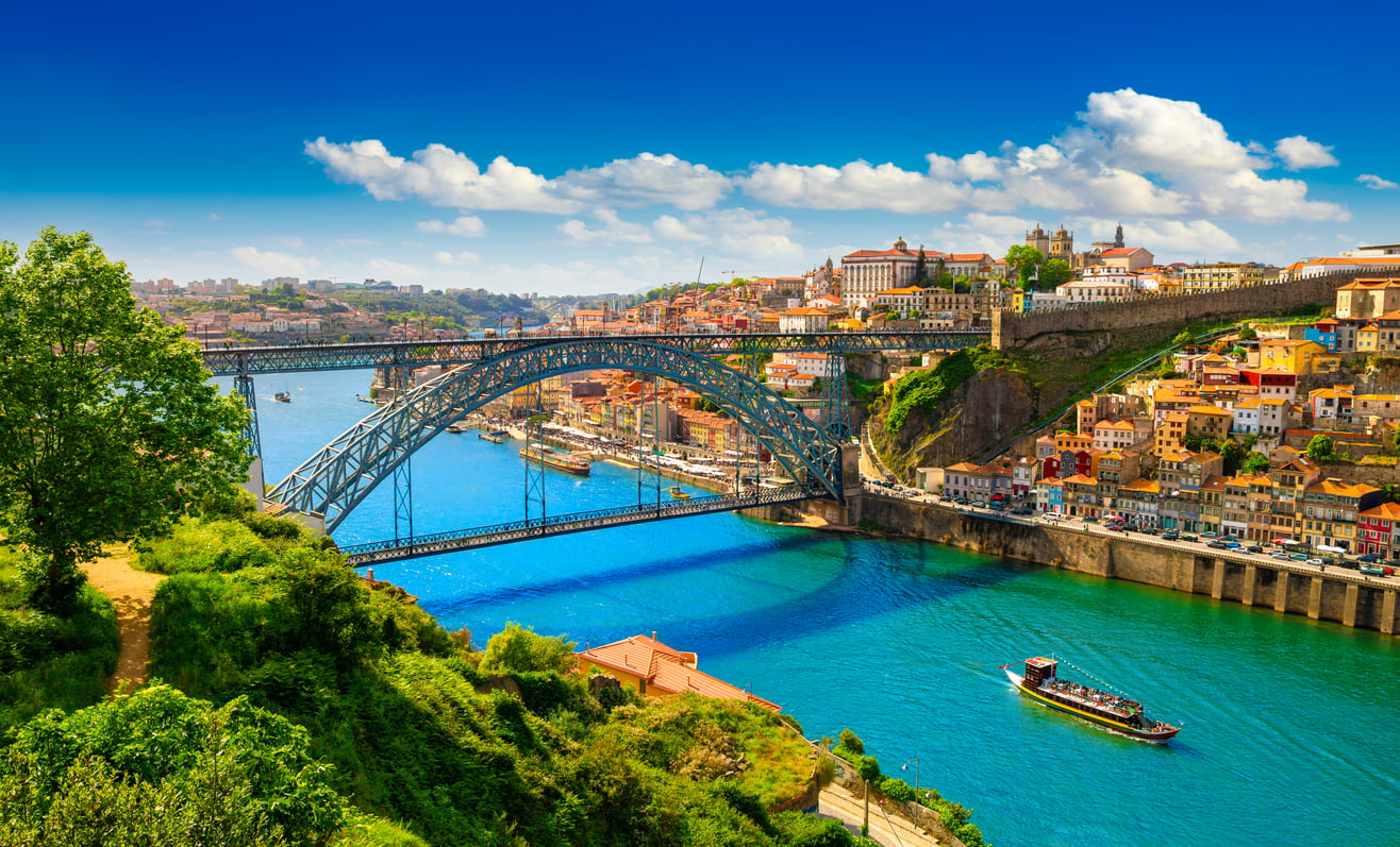 A view of a large arched metal bridge over a blue river, with a boat below, colorful hillside buildings, green trees, and a bright blue sky with scattered clouds—a picturesque scene from one of the travel spots boomers are learning to love.