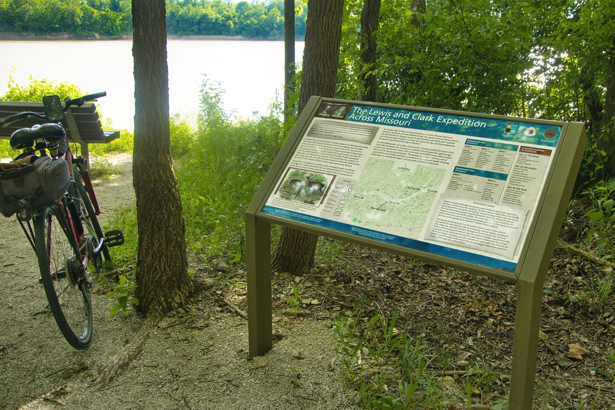 Bicycle parked near a trail sign titled “The Lewis and Clark Expedition Across Missouri,” with trees and a river in the background on a sunny day—a perfect spot to discover travel facts about the United States.