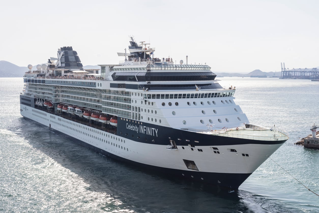 A large cruise ship named Celebrity Infinity sails near a port with cranes visible in the background. The ship is white with dark blue accents and is surrounded by calm water under a partly cloudy sky.