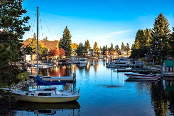A calm lake lined with docked boats and pine trees, with houses in the background under a clear blue sky, makes this one of the most serene vacation spots to relax and unwind.