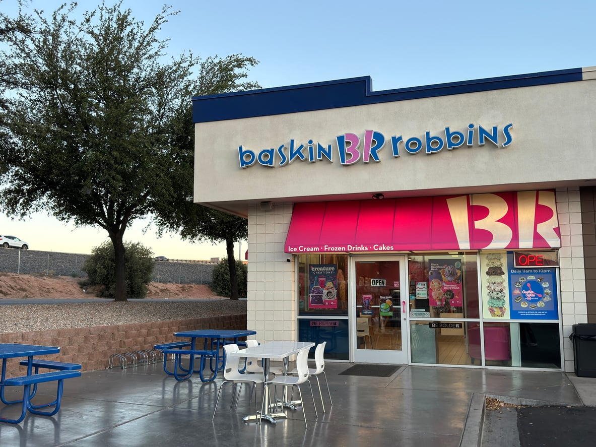 Baskin Robbins ice cream shop exterior with pink signage, blue picnic tables, a tree nearby, and clear sky in the background.