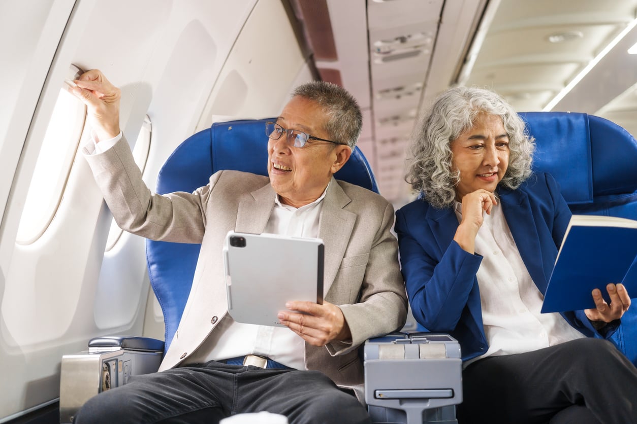 Two older adults sit on an airplane, demonstrating relaxed travel behaviors. The man holds a tablet and opens the window shade, while the woman beside him smiles and reads a book. Both appear comfortable, seated in blue airplane seats.