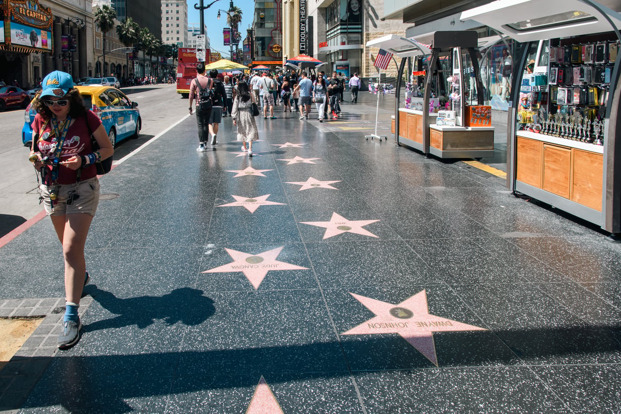 People walk along the Hollywood Walk of Fame, with souvenir stands and stars on the sidewalk, including Dwayne Johnson’s star, on a sunny day.