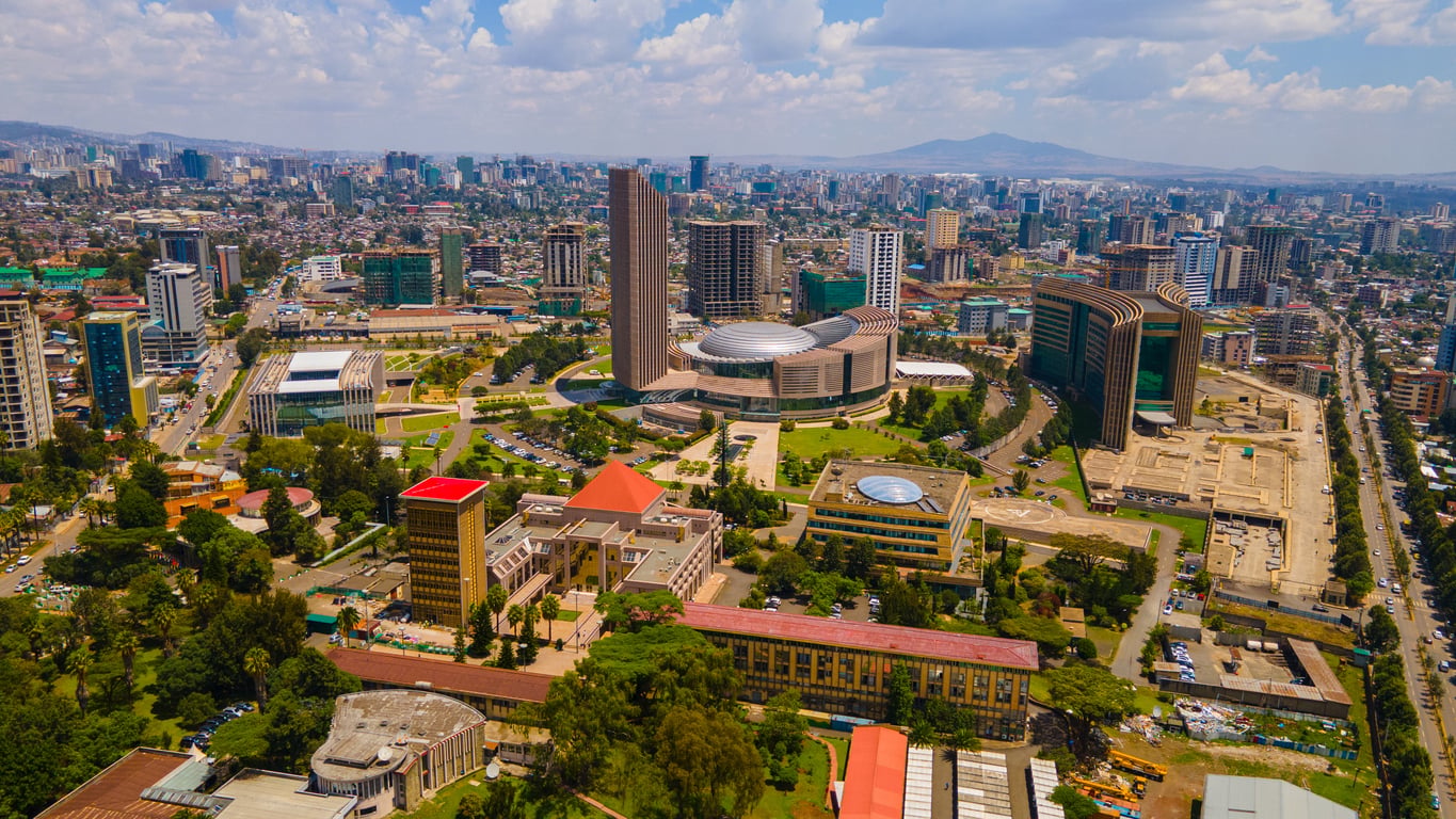 Aerial view of a modern city with tall buildings, green spaces, and a large round building in the center under a partly cloudy sky. Mountains are visible in the background, offering a picturesque destination for visitors—though it’s important to stay up-to-date on travel restrictions and advisories before planning your trip.