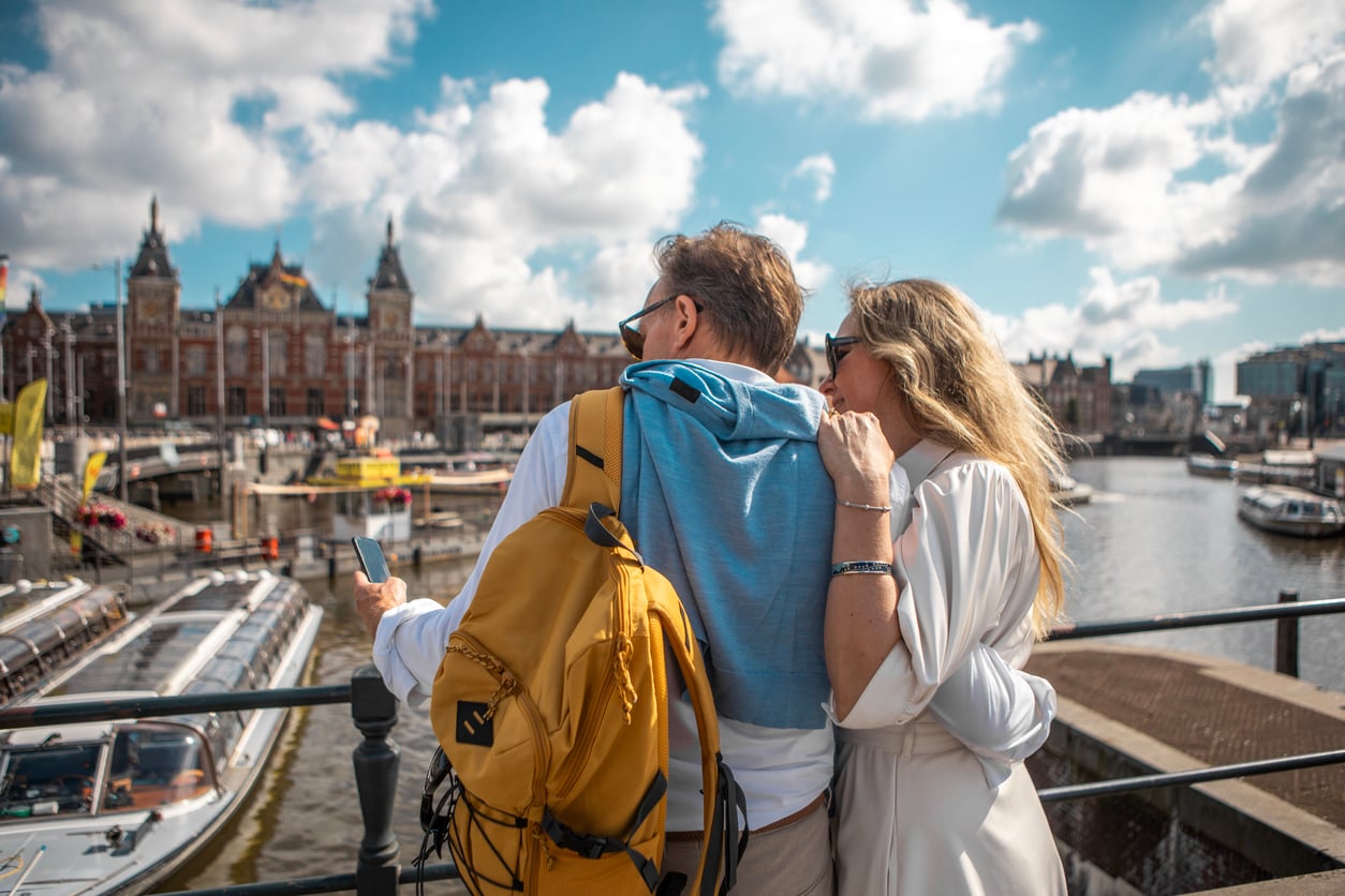 A couple stands by a canal, looking at a phone—perhaps searching for travel destinations Boomers are skipping. The man wears a yellow backpack and blue sweater, while the woman leans on his shoulder. Boats and a large building are visible across the water under a bright, partly cloudy sky.