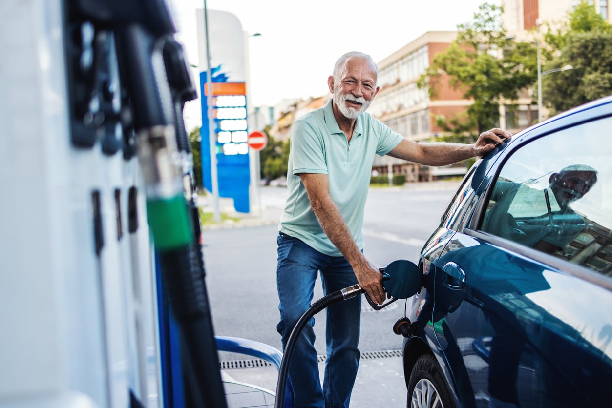 An older man with a white beard smiles while pumping gas into his car at a gas station on a sunny day. He is wearing a light green polo shirt and jeans. The gas station and street are visible in the background.