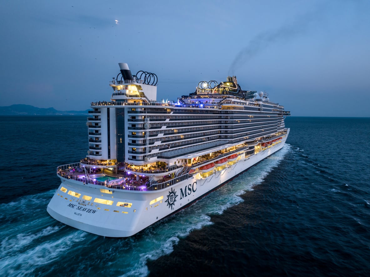A large white MSC cruise ship sails on calm blue water at sunset, with lights glowing on multiple decks and passengers visible outdoors.