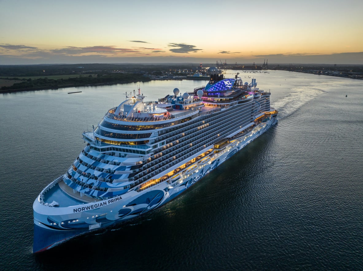 A large, modern cruise ship named Norwegian Prima, part of one of the world’s premier cruise lines, sails on calm water at sunset, illuminated by lights. The coastline and distant city skyline are visible in the background.