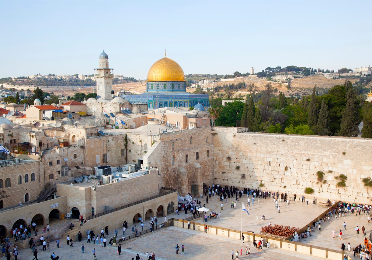 View of Jerusalem’s Western Wall with people praying, the Dome of the Rock with its golden dome in the background, and city buildings under a clear sky.