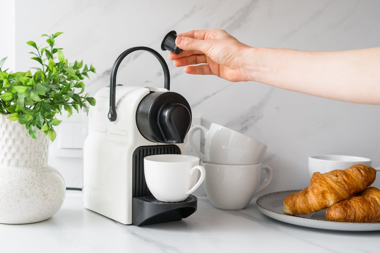 A hand places a coffee capsule into a white coffee machine with a cup underneath. Nearby are stacked white cups, a plate with croissants, and a potted plant on a white countertop.