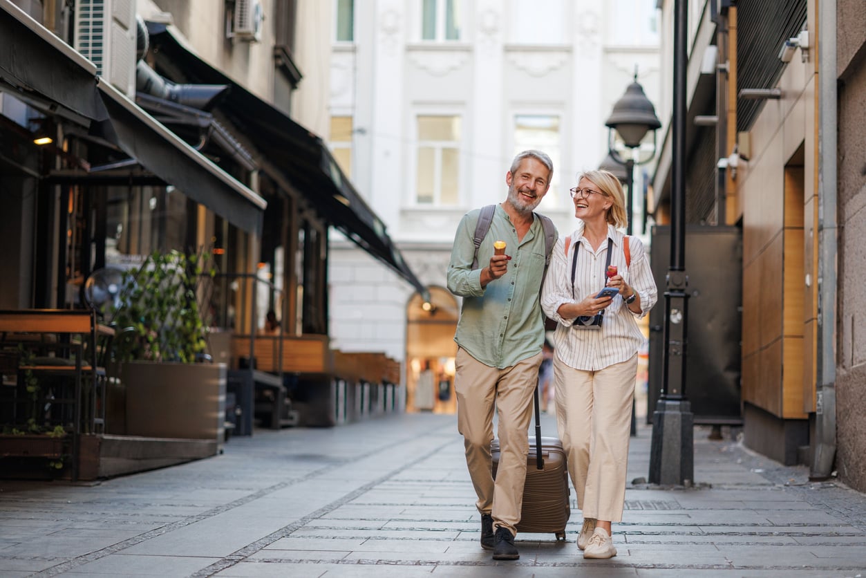 An older couple walks down a city street, smiling and eating ice cream, their travel habits on display with a rolling suitcase and a backpack, surrounded by outdoor restaurants and tall buildings.