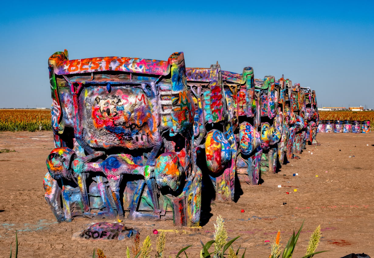 Ten partially buried cars, covered in colorful graffiti, stand in a row in a dirt field under a clear blue sky at Cadillac Ranch in Amarillo, Texas—one of the most iconic Classic Roadside Attractions in America.