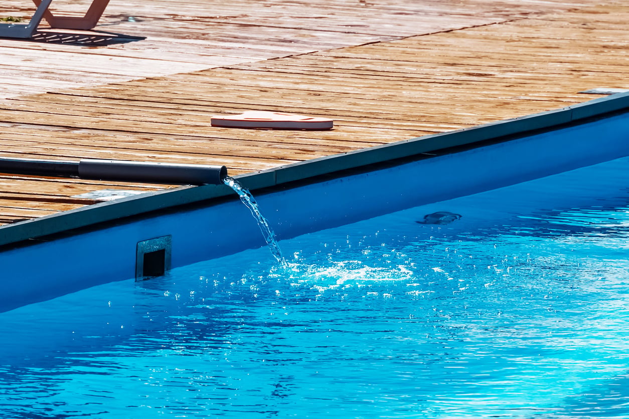 Water flows from a black pipe into a bright blue swimming pool next to a wooden deck. A pink object lies on the deck in the sunlight.