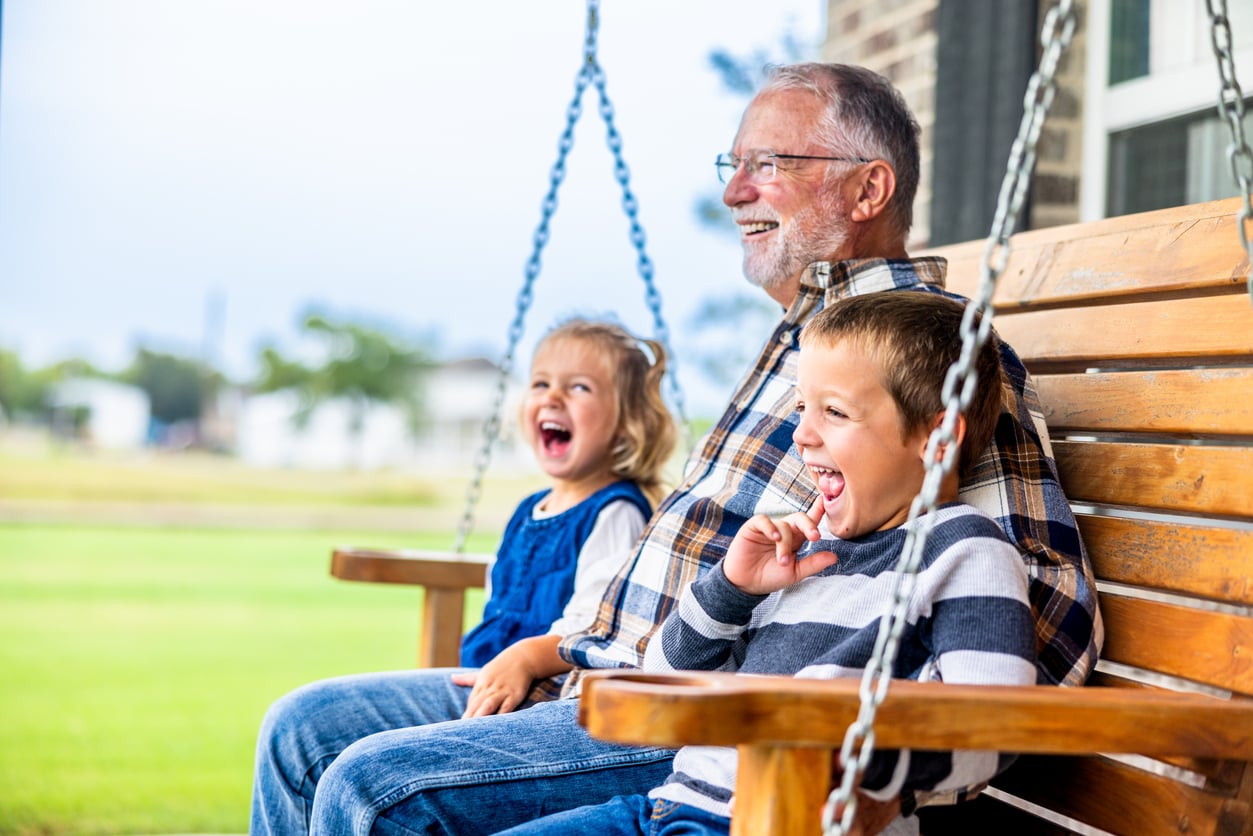An older man and two young children laugh together while sitting on a wooden porch swing outside on a bright day, capturing the warmth and joy that reflect the positive influence of Baby Boomer changes on family life.