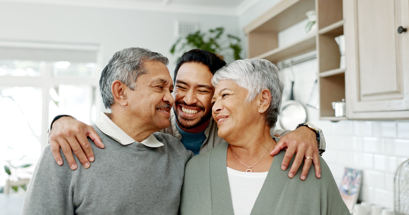 A smiling adult man hugs and stands between two older adults in a cozy kitchen, all three appearing happy and content together—capturing a warm moment that reflects the positive impact of Baby Boomer changes on family bonds.