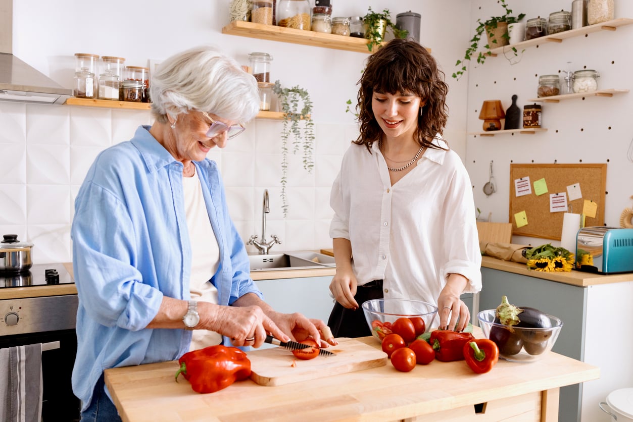 Two women prepare vegetables together in a bright kitchen. One chops a red bell pepper while the other smiles, surrounded by tomatoes and eggplants on the wooden counter.