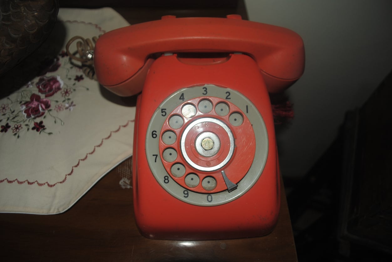 A vintage red rotary dial telephone sits on a wooden table next to an embroidered white cloth with floral patterns.