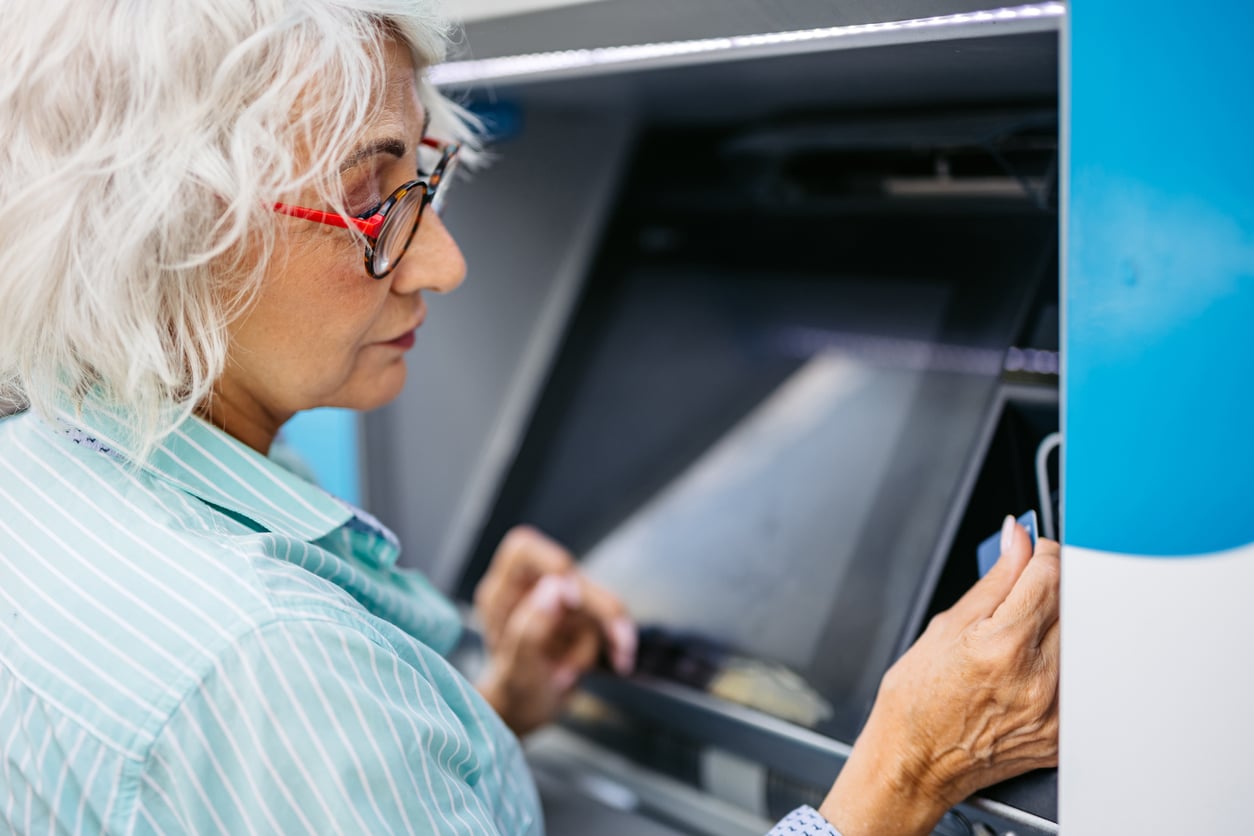 An older woman with gray hair and glasses uses an ATM, holding a bank card near the card slot and looking at the screen, perhaps considering some helpful retirement tips as she manages her finances.