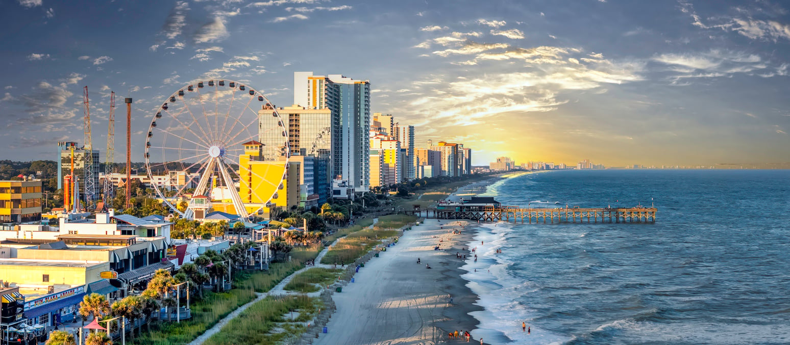 A panoramic view of a beachfront city, one of the most popular vacation spots, with tall hotels, a Ferris wheel, a pier extending over the ocean, and people walking along the sandy shore at sunset.