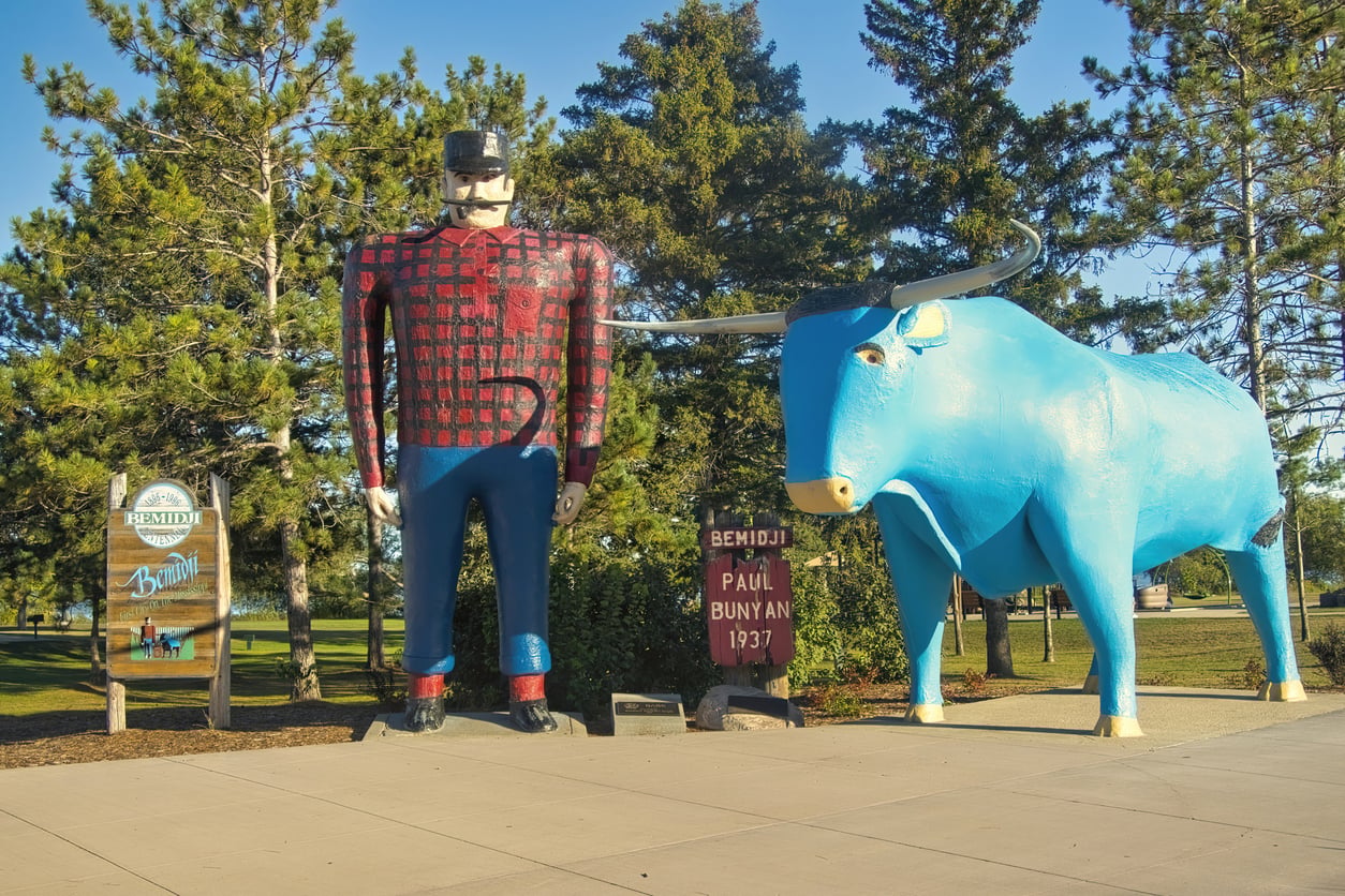 Statues of a lumberjack in red plaid and a large blue ox stand on a sidewalk surrounded by trees, creating one of the region’s Classic Roadside Attractions, with signs nearby reading “Bemidji” and “Paul Bunyan 1937.”