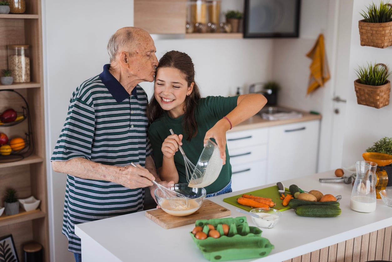 An elderly man and a young woman smile while cooking together in a kitchen, mixing ingredients in a bowl surrounded by fresh vegetables, eggs, and milk.