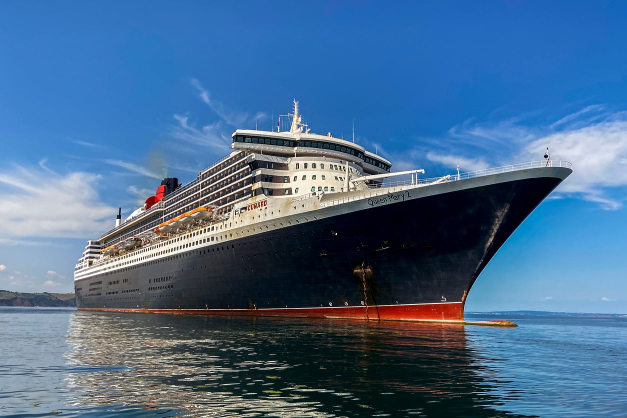 A large black and white cruise ship with multiple decks sails on calm water under a blue sky, with “Queen Mary 2” visible on its bow.