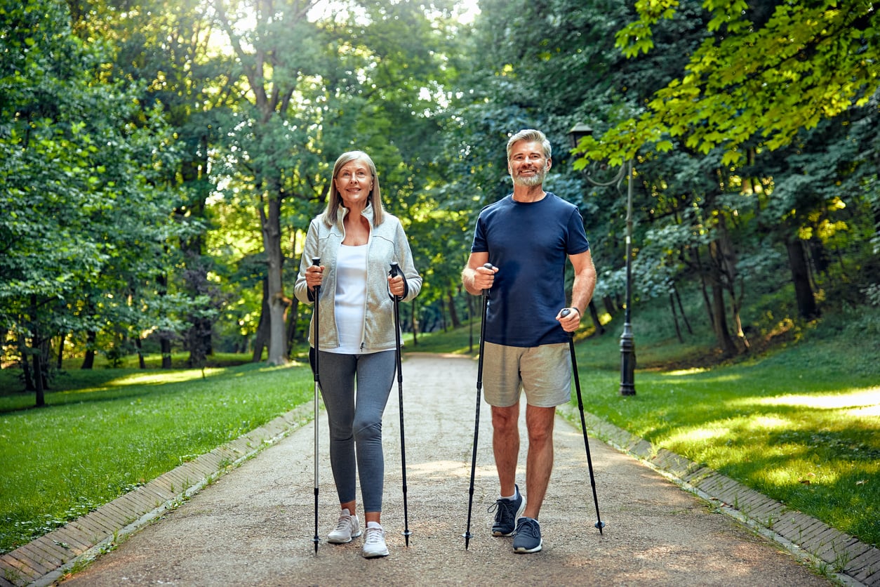 Two adults smiling and Nordic walking on a tree-lined park path, holding walking poles, with green grass and sunlight filtering through the trees around them.