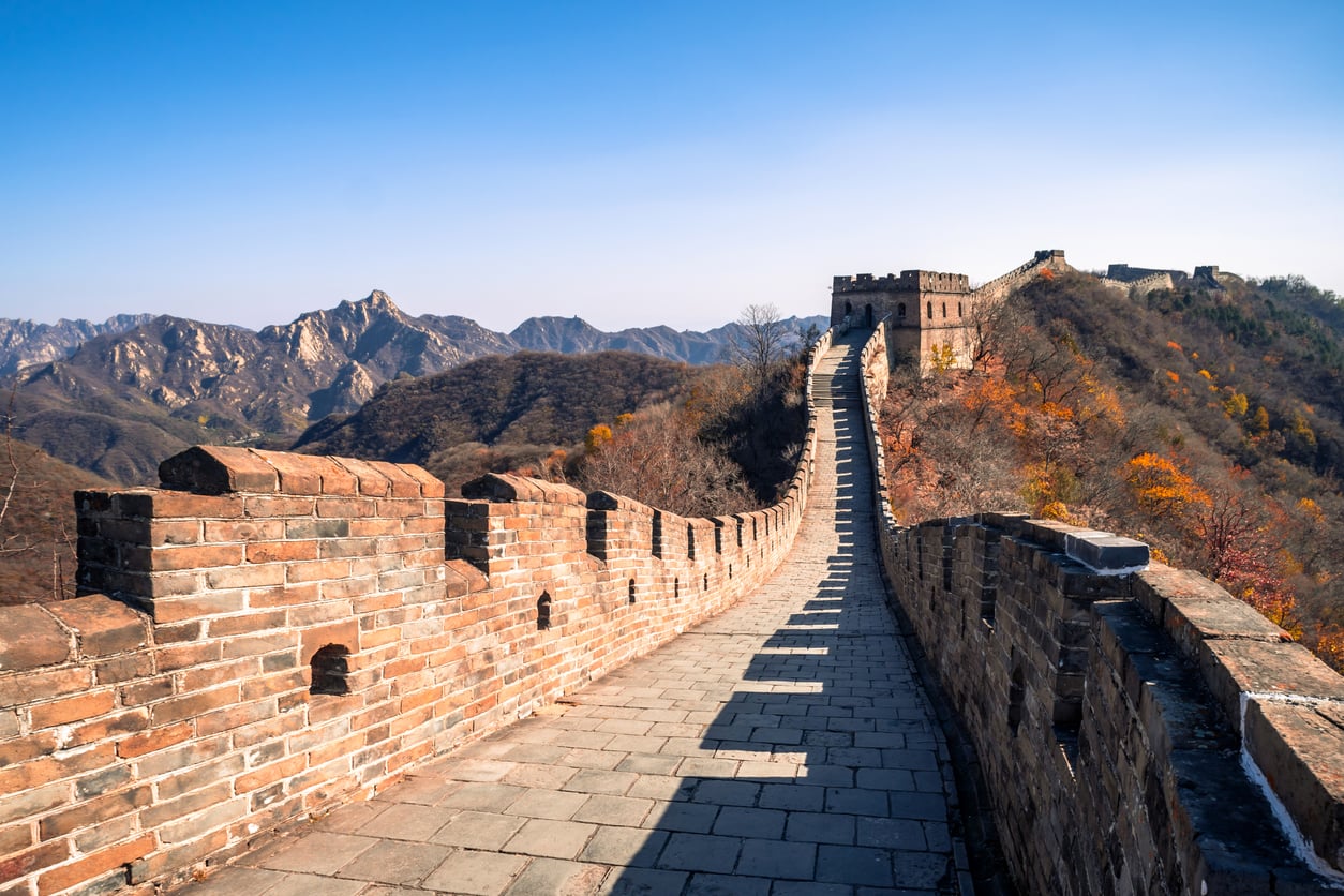 A section of the Great Wall of China winds over hills under a clear blue sky, with watchtowers and mountains in the background.