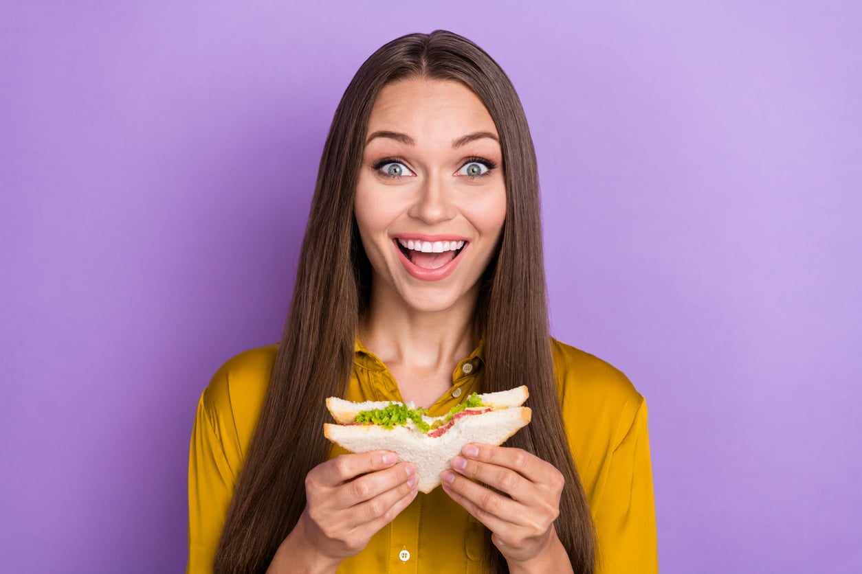 Smiling woman with long brown hair in a yellow shirt holds a sandwich with lettuce and tomato, standing in front of a purple background.