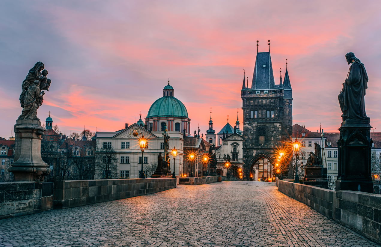 Charles Bridge in Prague at sunrise, with streetlights glowing, statues lining the sides, cobblestone pavement, and the Old Town Bridge Tower in the background under a colorful sky, is fast becoming one of those travel spots Boomers are learning to love.